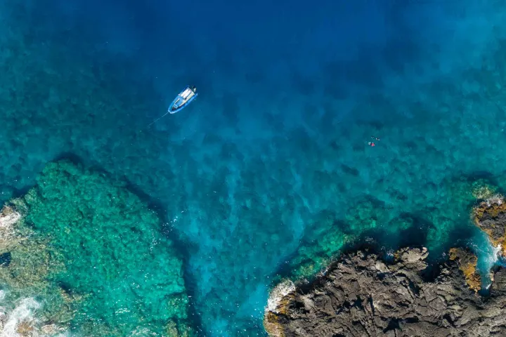 overhead view of a snorkel boat and people snorkeling near a lava rock coastline in tropical waters