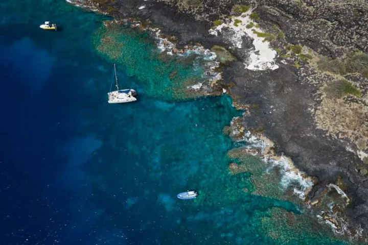 view of Kona's rugged coastline from the air with 2 boats sitting in the water near shore and a reef below