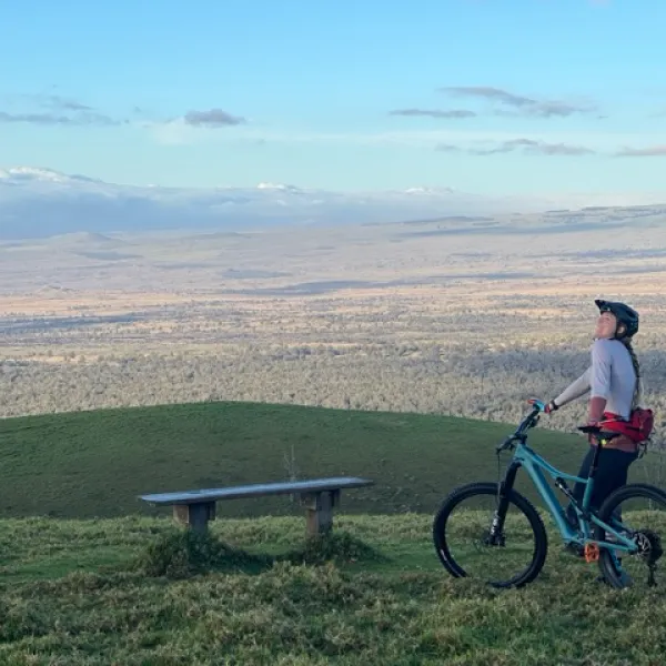woman on a mountain bike posing awkwardly on a grassy hill with yellow dead grassy view in the background and clouds on mountains behind