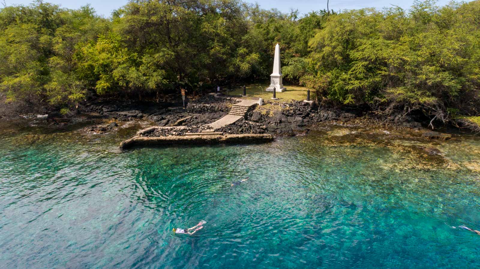 a snorkeler swims in tropical shallow water near the shore where a white obelisk sits among the trees