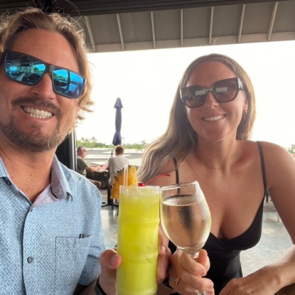 a woman wearing glasses holding a glass of wine cheersing with a man wearing sunglasses and holding a birght colored tropical drink
