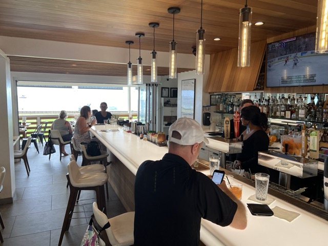 a man sitting at a brightly lit bar with a bartender and a view out to the restaurant interior and view outside of the ocean