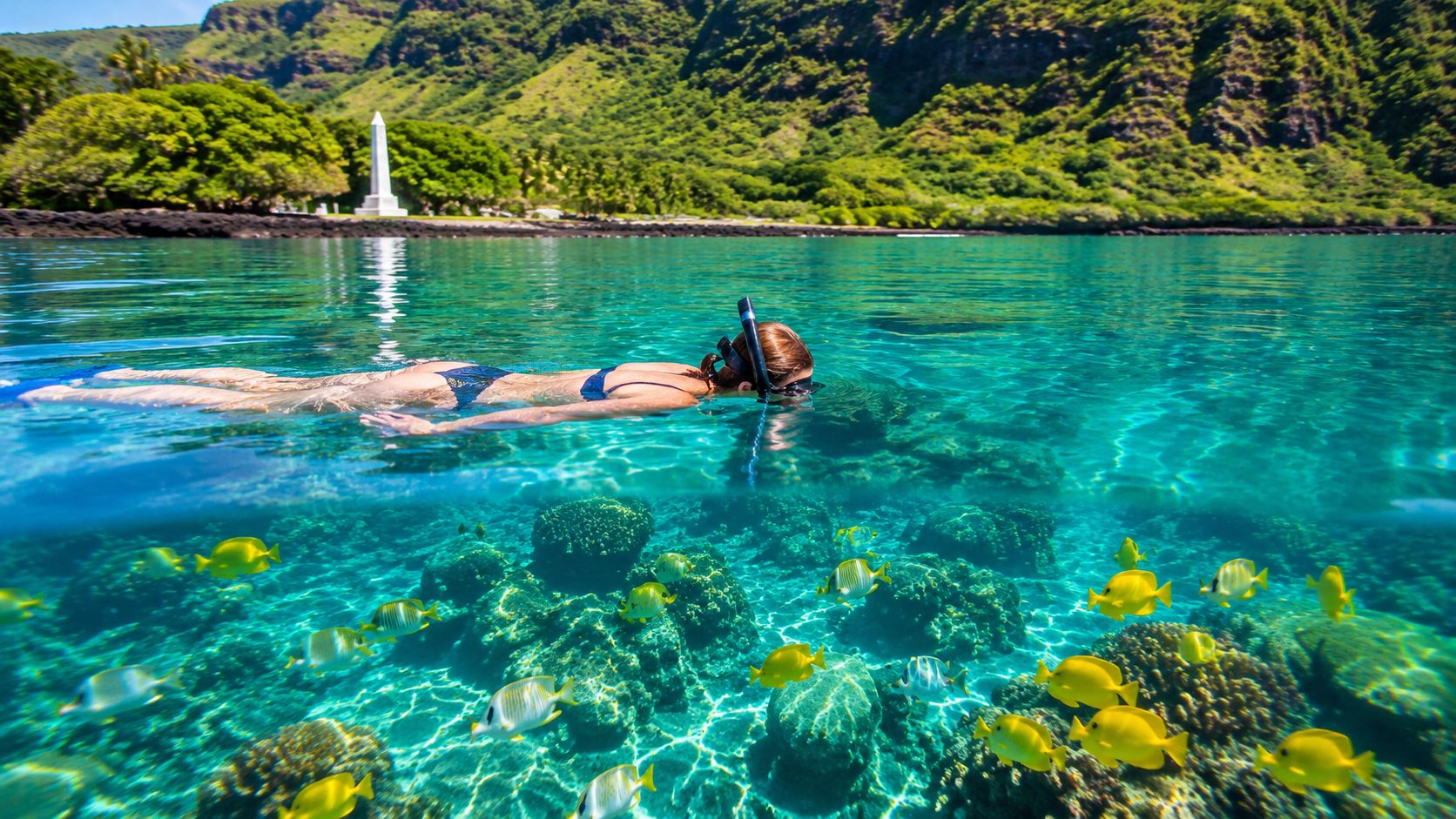 Person snorkeling over coral reef with yellow fish in clear blue water, lush green mountains in the background.
