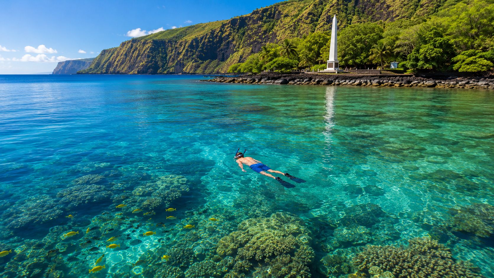 Person snorkeling near coral reef in clear blue water, with a monument and lush cliffs in the background.